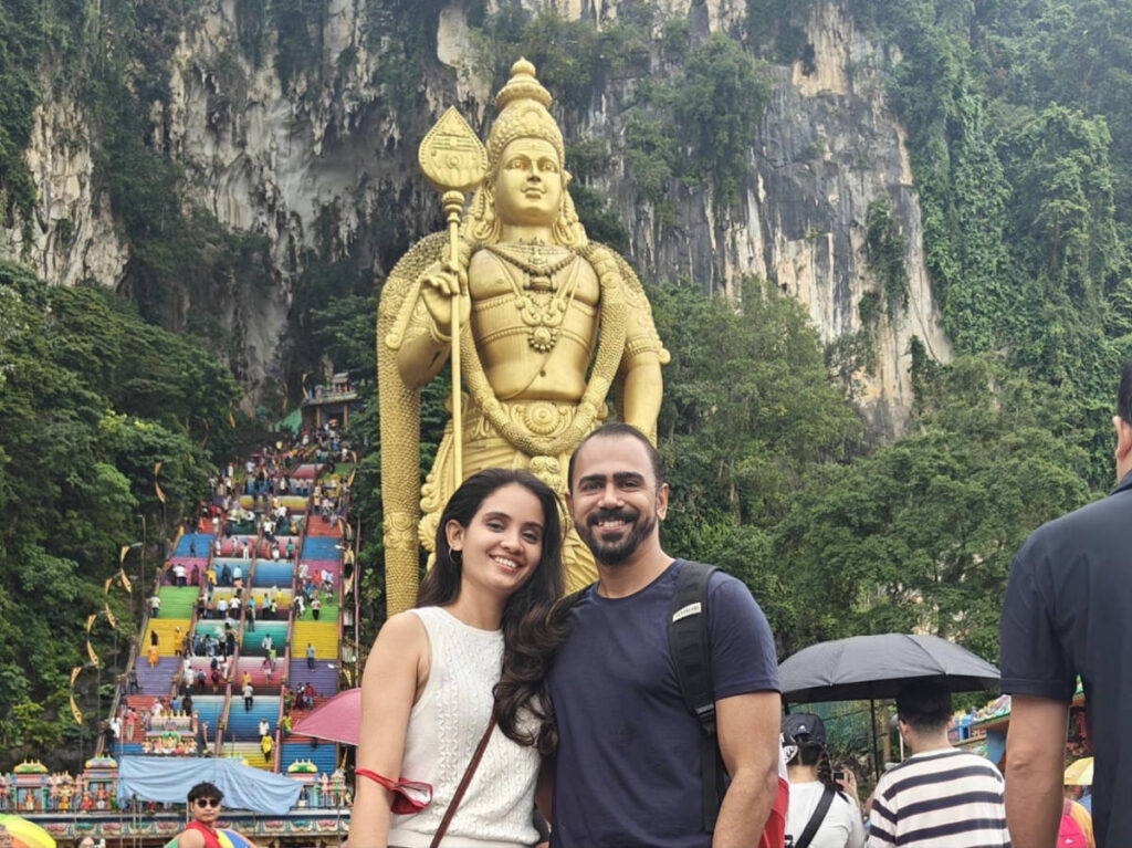 The author, Advaita and her husband, smiling for a photo with the colorful staircase and statue of Lord Murugan in the background