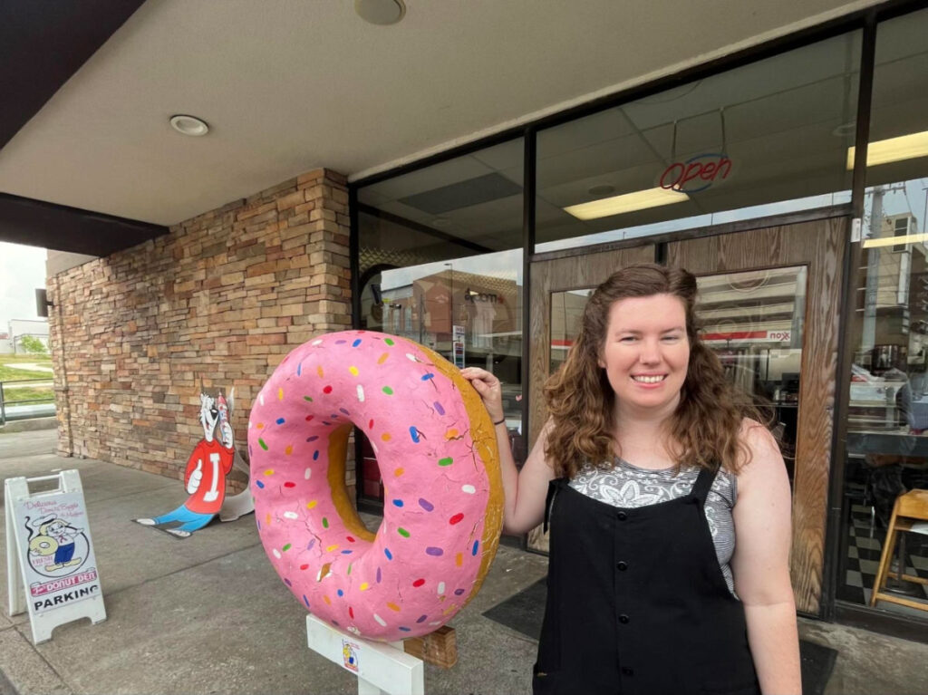 Author Betty posing for a photo with a large donut outside Fox’s Donut Den