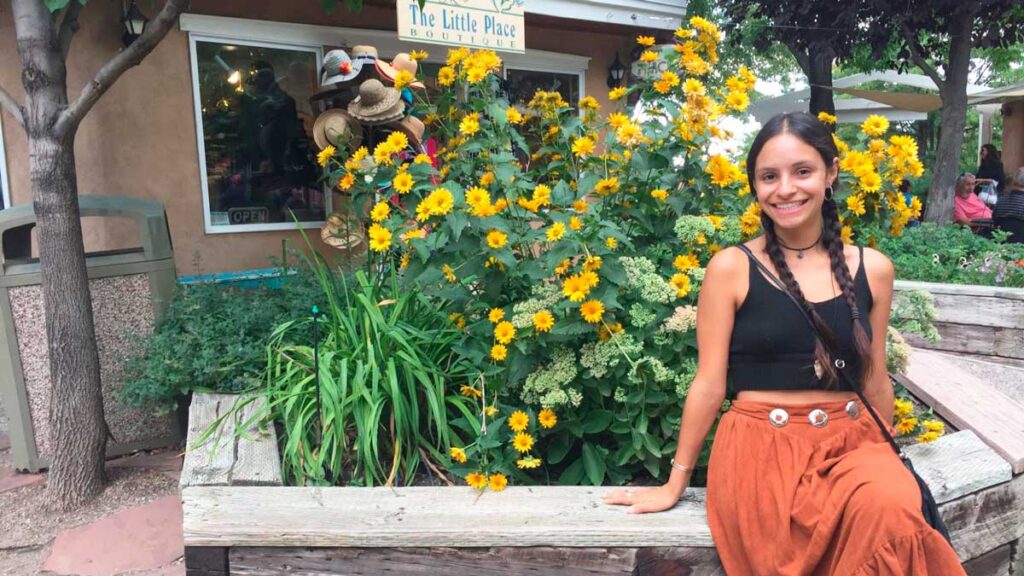 The author McKenna Mobley smiling for a photo in Taos Plaza with colorful flowers on her background