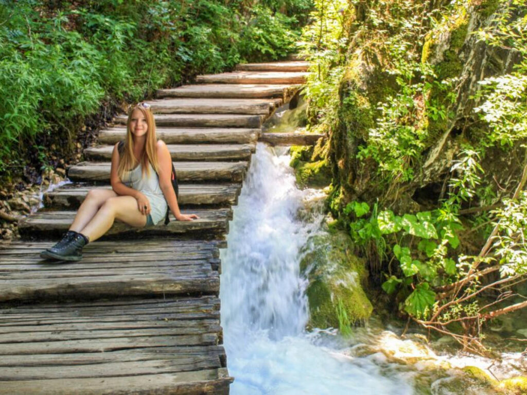 Author Taylor sitting on a wooden stairs near a waterfall in Plitvice Lakes National Park
