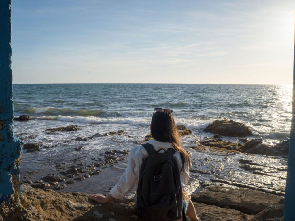 Author Vanessa with a backpack sitting on rocks by the sea 