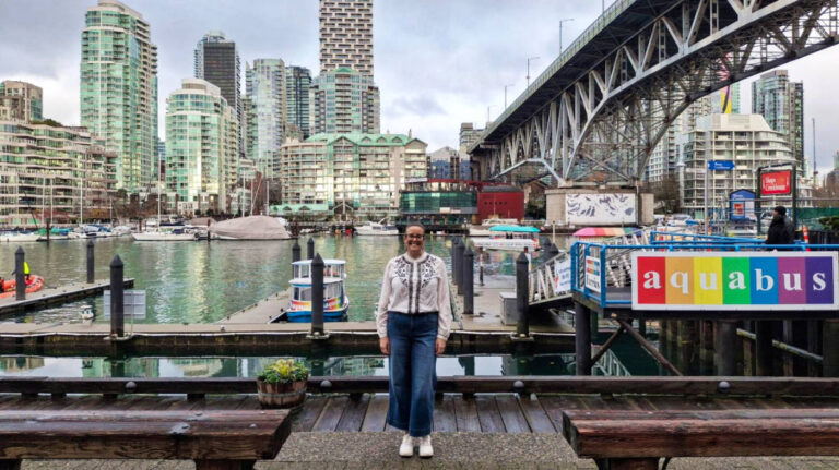 The author Catherine Driver, smiling for a photo at the Granville Island with city views in the background