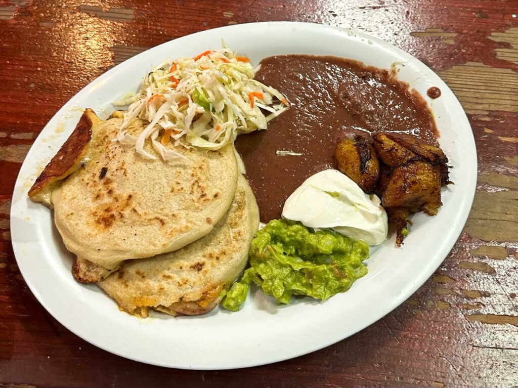 A plate with chicken pupusas, beans, sour cream, guacamole, sweet plantains, and coleslaw