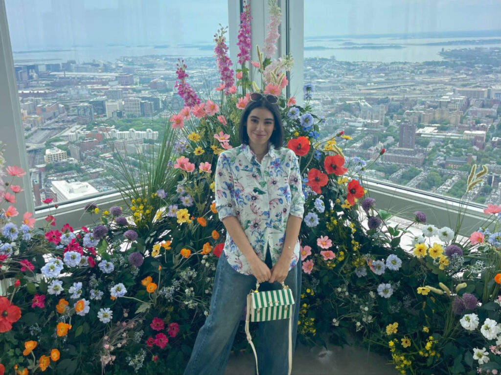 The author Claudicet Pena, smiling for a photo with flowers around with an overlooking view of Boston