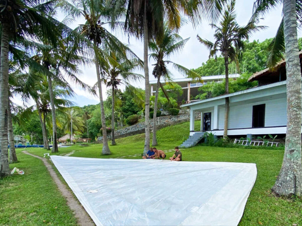 The sailing crew patching up a hole on the mainsail under the coconut trees
