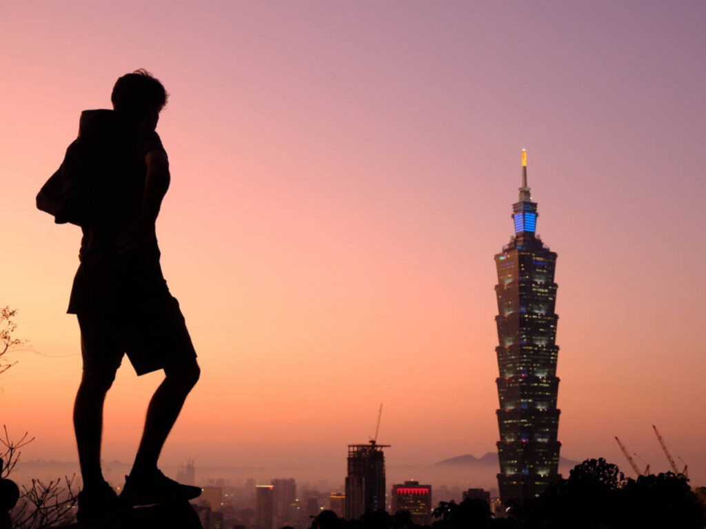 A man's silhouette from Elephant Mountain and the Taipei 101 building from afar during sunset