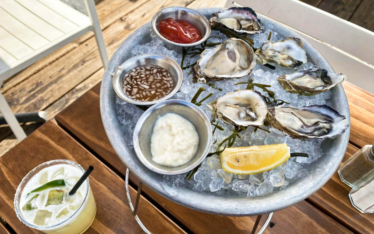 Fresh oysters with different sauces on the side served on a tray with ice and a glass of drink