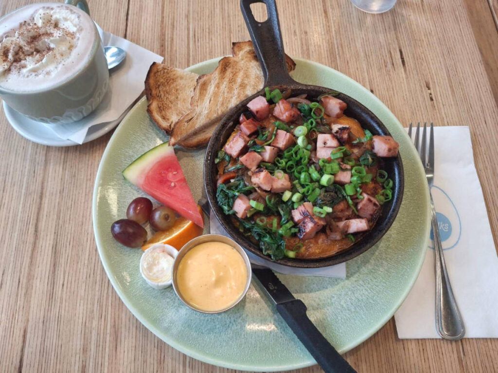 A huge breakfast plate with Fri-tot-a on a skillet pan, toasted breads, fruits, sauce and a glass of drink on the side