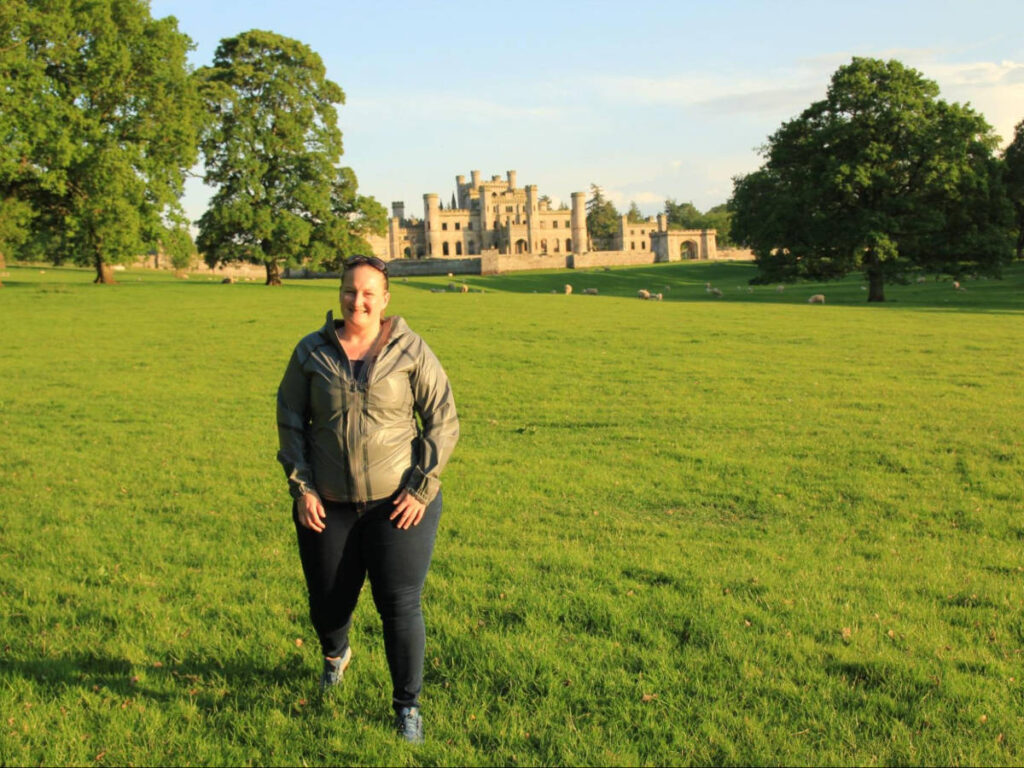 The author Heather Cole, posing for a photo with the view of Lowther Castle surrounded by greenery