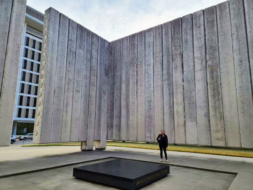 Author Sarah Heath standing in front of a large concrete wall at John F. Kennedy Memorial Plaza
