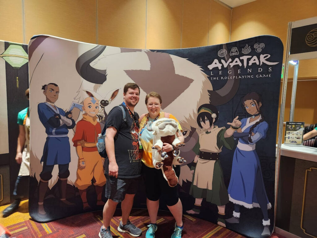 The author Kelsey Nighthawk and her husband posing in front of the Avatar Legends Game booth during Gen Con