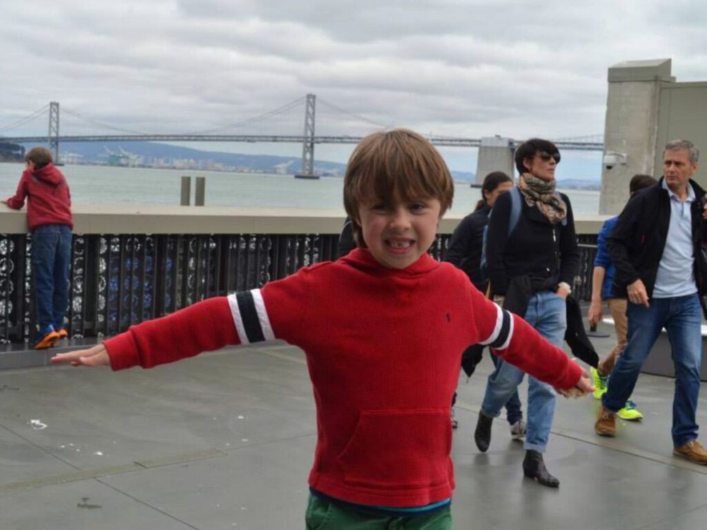 A young boy with arms outstretched on a sidewalk with the San Francisco bridge in the background