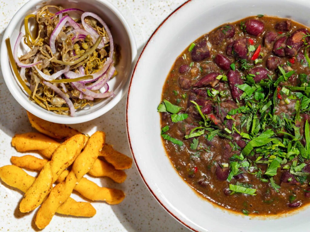 A bowl of red bean dish with breadsticks and pickled onions on the side