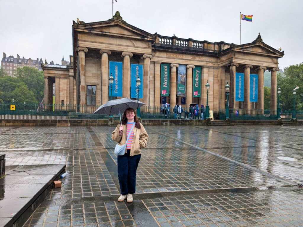 The author Louise Wylie smiling for a photo in front of The National Gallery of Scotland on a rainy day
