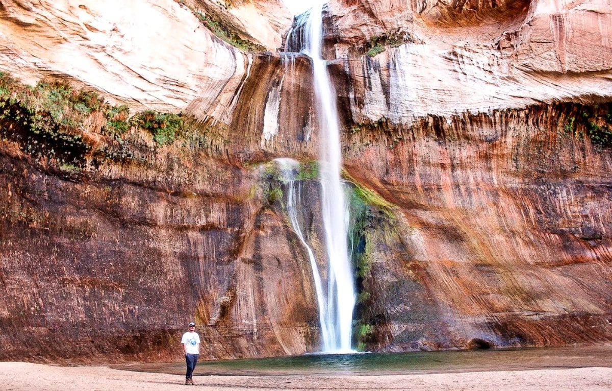A man standing near the tall waterfalls in The Lower Calf Creek Falls Trail