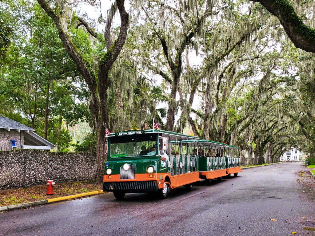 A green and orange trolley moving along Old Town St. Augustine with trees on both sides