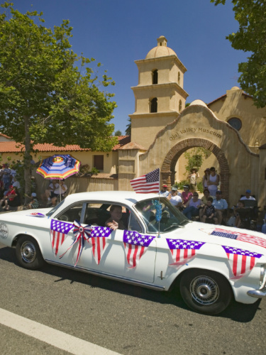A white car adorned with decorations drives in a parade in front of Ojai Valley Museum