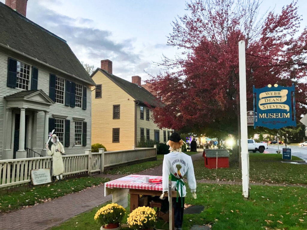 A huge red tree, signage, scarecrow near a table with flowers outside the Webb Deane Stevens Museum