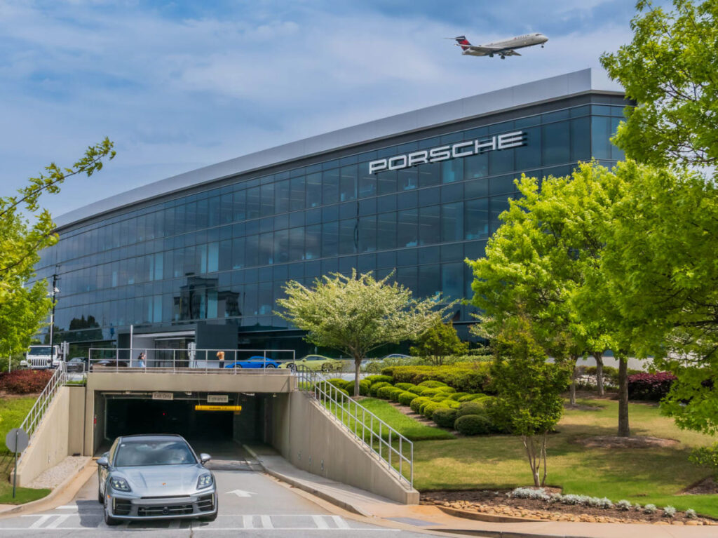 A Porsche drives out from the Porsche Experience Center, and an airplane flying low overhead