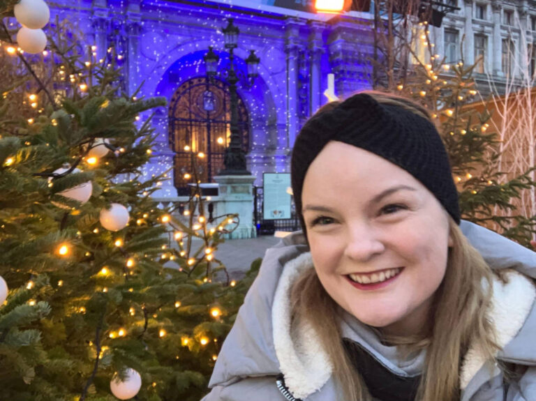 The author Rachel Kapelke-Dale, smiling in front of the decorated Christmas trees at the Paris’ City Hall Christmas Village