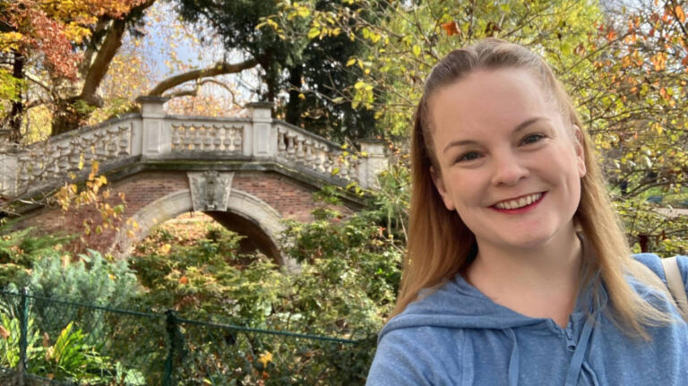 The author Rachel Kapelke-Dale, smiling for a photo in front of the bridge at the Parc Monceau