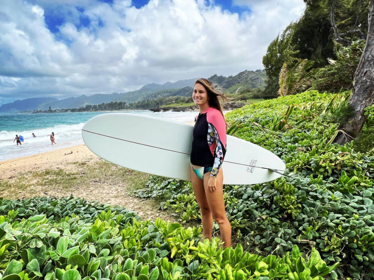 The author Rebecca Parsons carrying her surfboard at the beach