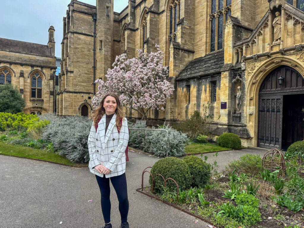 The author Rebecca Rayner, posing for a phot o outside the Mansfield College with cherry blossoms in front