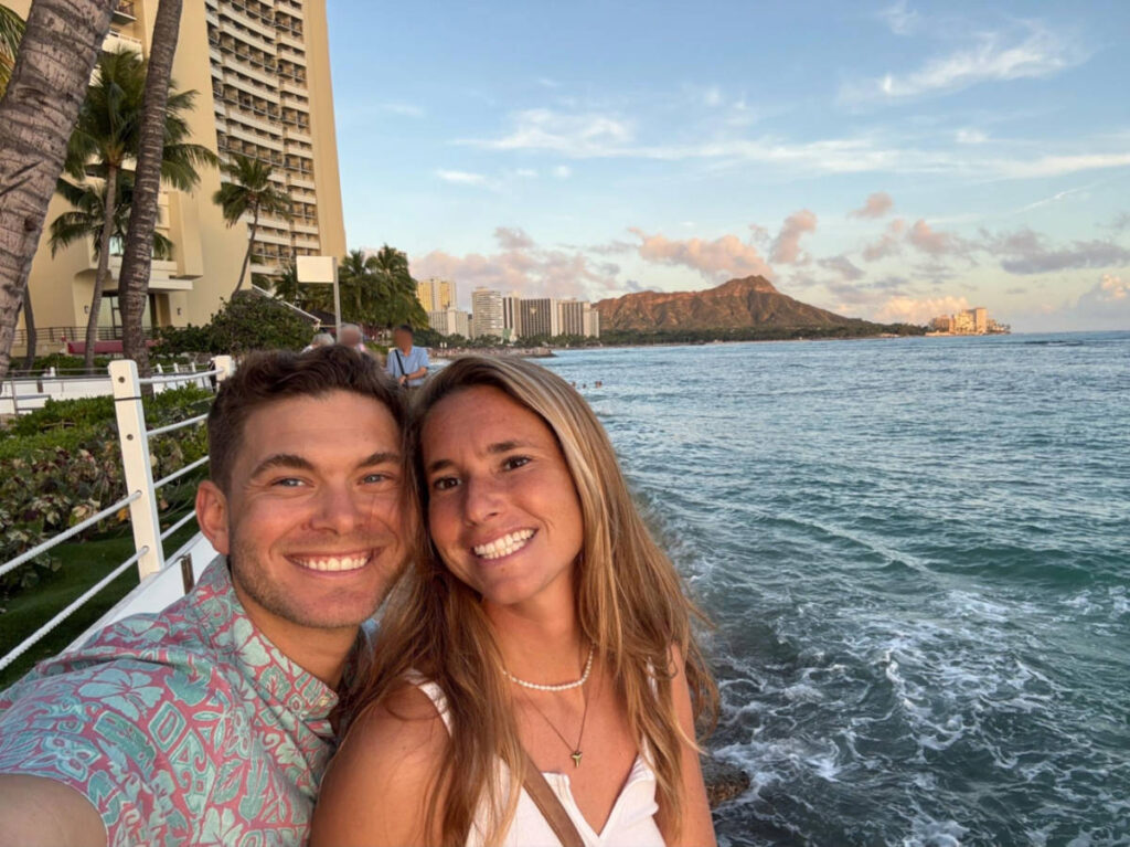 The author Rebecca Parsons and her partner smiling for a photo with the scenic view of Waikiki