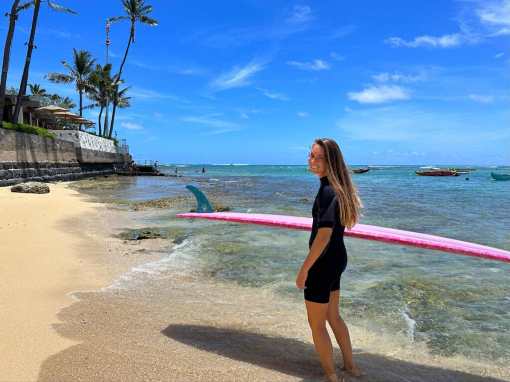 The author Rebecca Parsons holding a surfboard along the shoreline of Oahu’s Gold Coast
