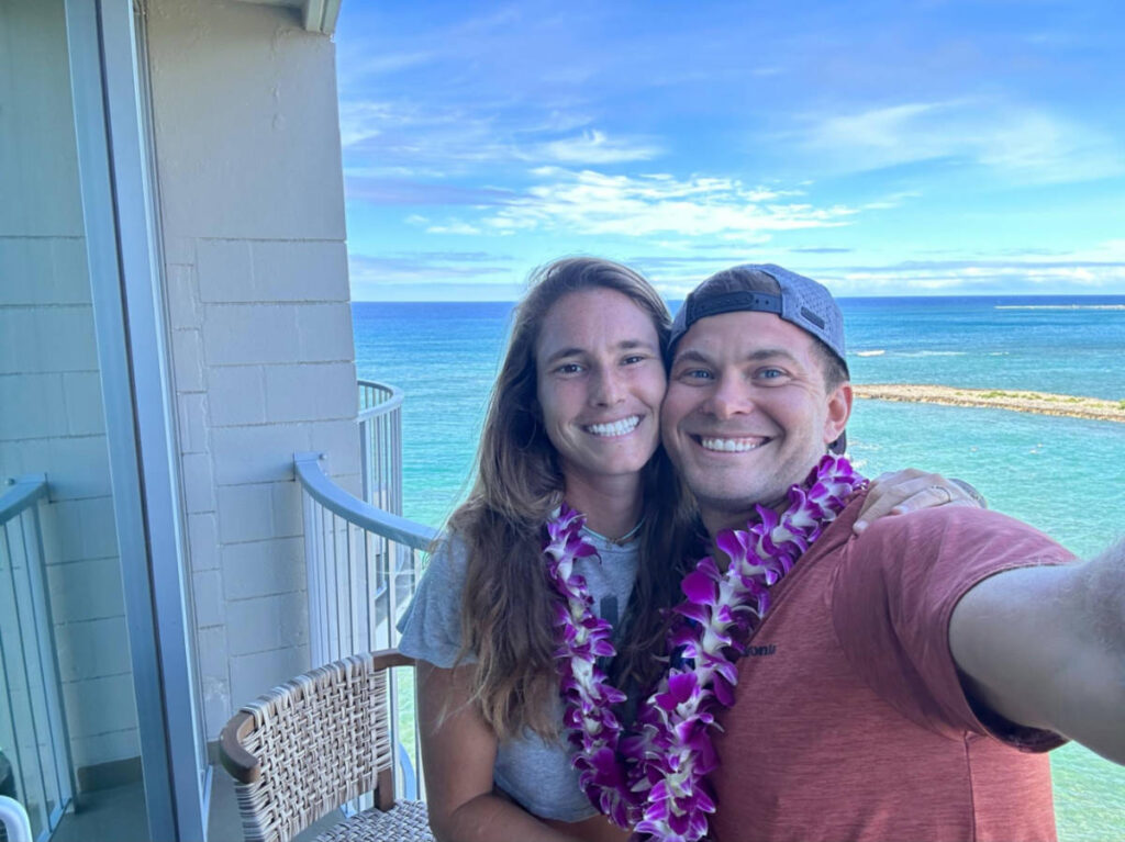 A couple smiling for a photo at the lanai of their hotel room at The Ritz-Carlton O'ahu