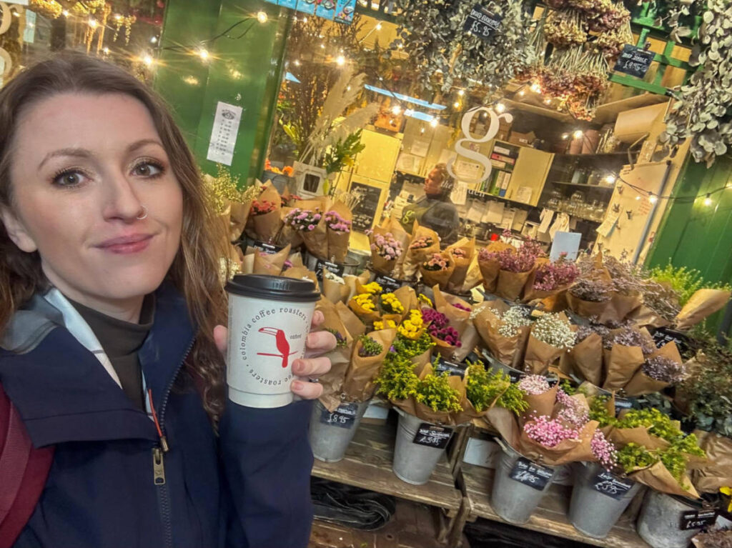The author Rebecca Rayner, holding a cup of coffee in front of the flower shop