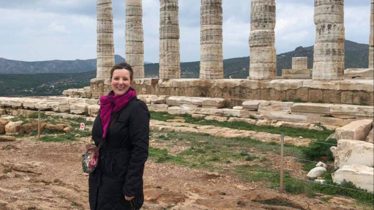 The author Rebecca Hall, smiling for a photo in front of the Temple of Poseidon at Cape Sounion