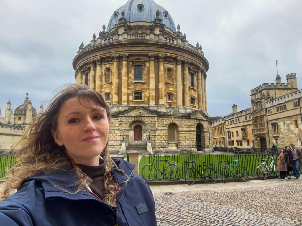 The author Rebecca Rayner, smiling for a photo in front of the historical landmark in Oxford on a gloomy day