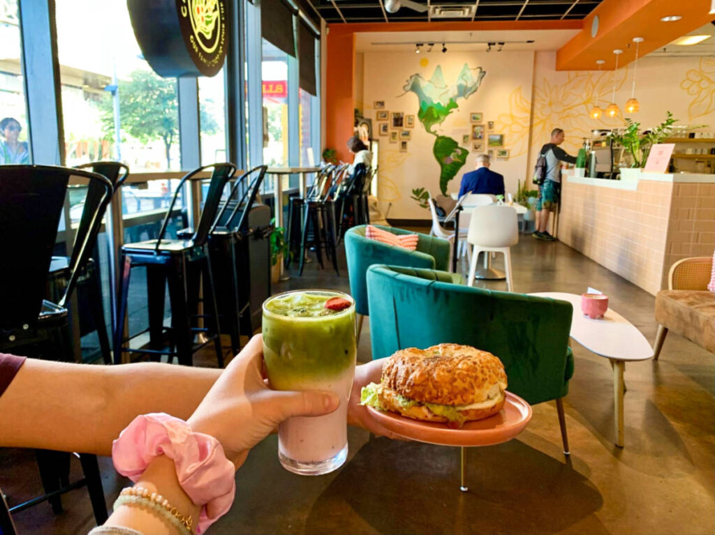 Hands holding a glass of rose matcha and a plate breakfast burger inside Cafetal Cafe