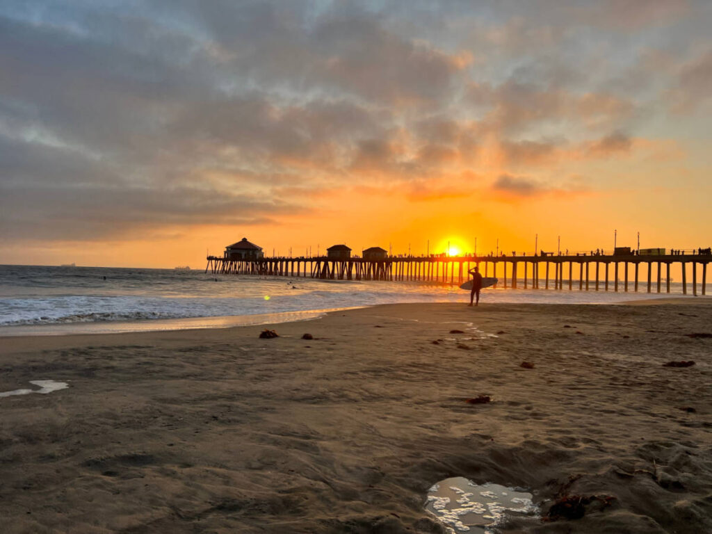A surfer walking near the Huntington Beach Pier at sunset
