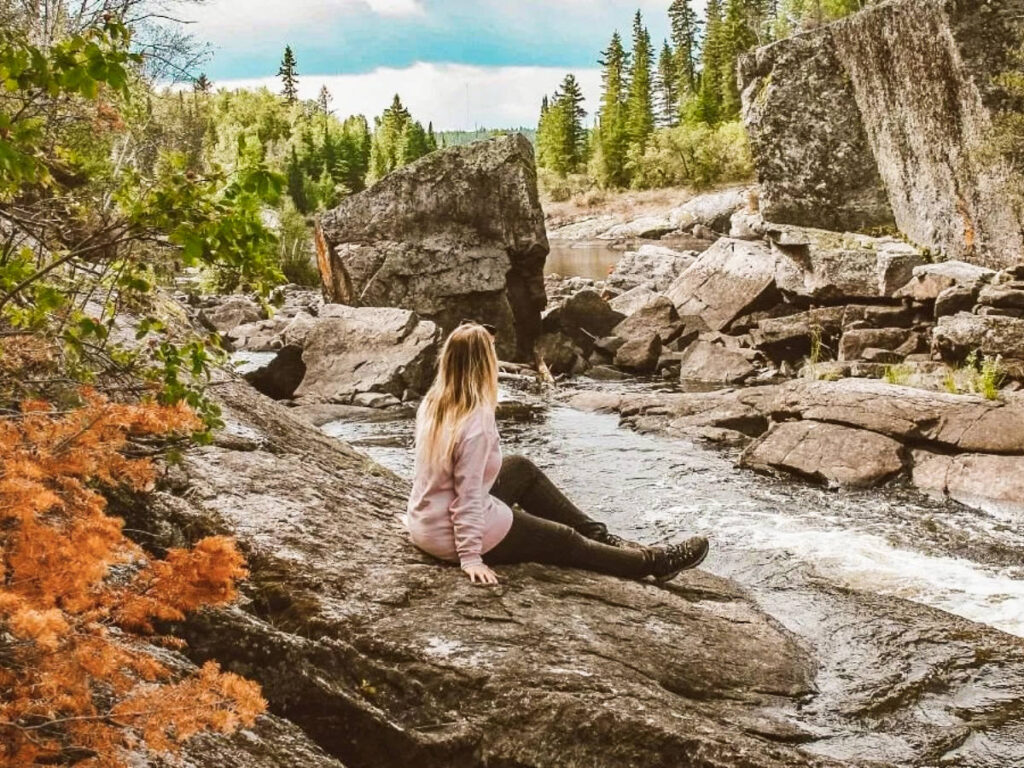 The author Taylor Herperger, sitting on the rock with the fall foliage around Tulabi Falls in Nopiming Provincial Park