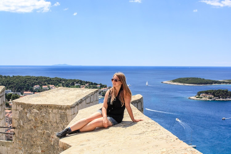 Taylor sits on the walls of the Spanjola Fortress in Hvar, with the Adriatic Sea, islands, and sailboats in the background.