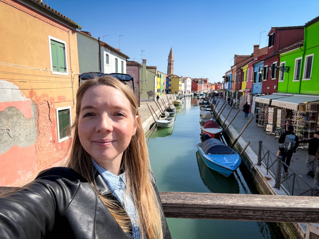 The author Taylor Herperger, smiling for a photo in front of the colorful buildings and the boats on the canal in Burano