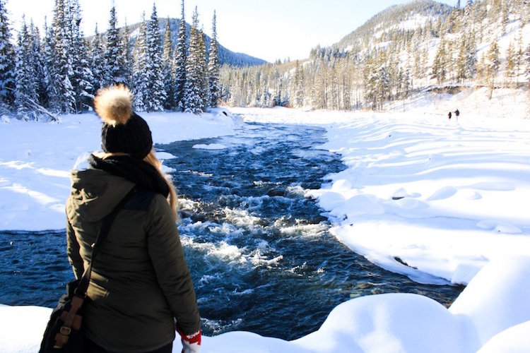 Taylor stands with her back to the camera as she looks out over a rushing river surrounded by powdery white snow, trees, and mountains in Kananaskis, Alberta