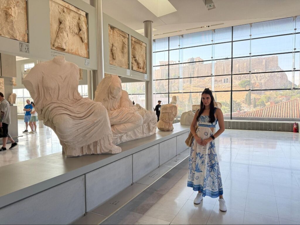 Travel Lemming creator Clara inside the Acropolis Museum, with the Acropolis visible through the window in the background