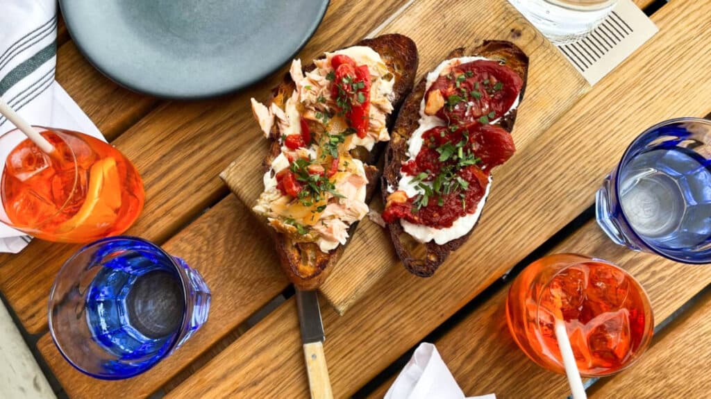 Two bruschetta topped with poached eggs and sliced tomatoes served on a wooden board with glasses of water and iced drinks around