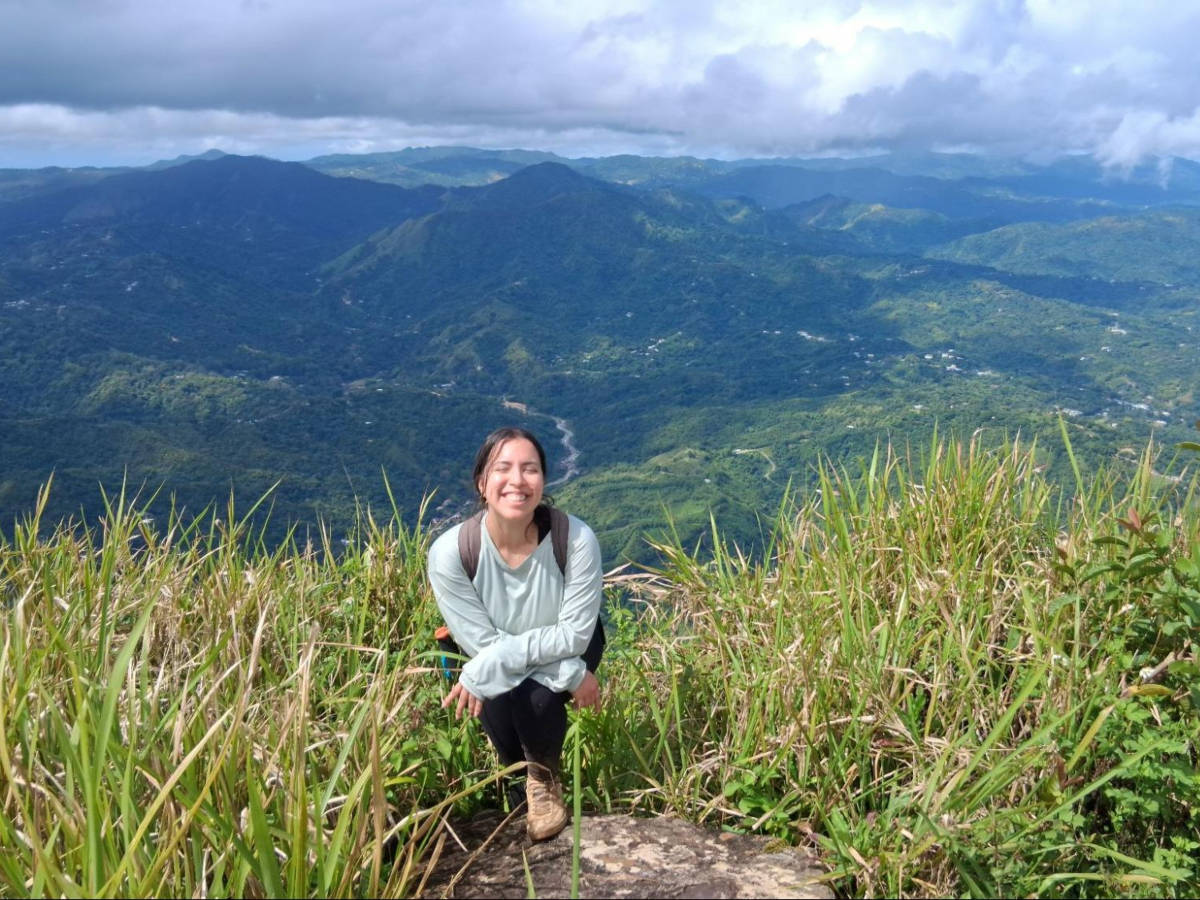 The author Vanessa Ramos, smiling for a photo at Cerro Morales Peak with a scenic overlooking view in the background