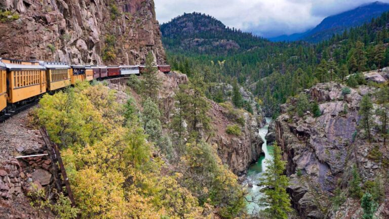 View of a yellow vintage train along the Durango and Silverton Narrow Gauge Railroad with a view of the mountains and river below