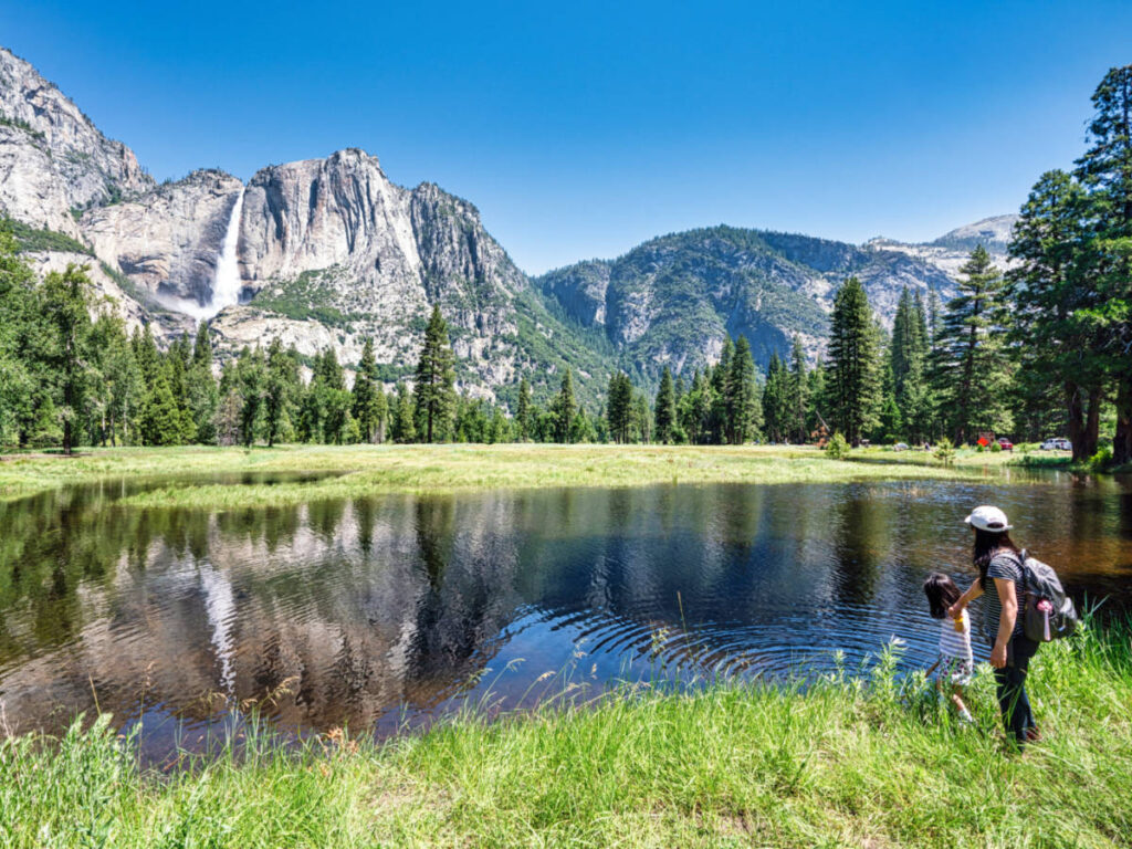 A woman and a kid hiking near the lake with the reflection of the falls on the mountain ranges